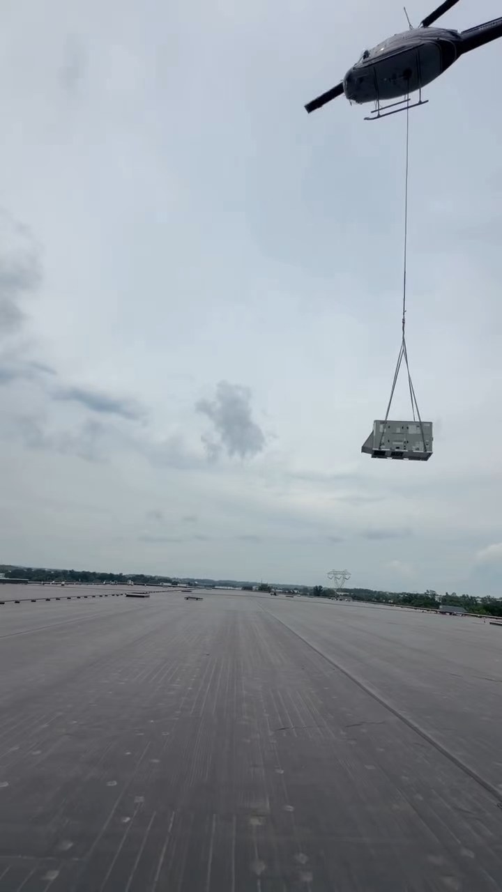Helicopter flying a rooftop HVAC unit across a large warehouse roof toward the set point