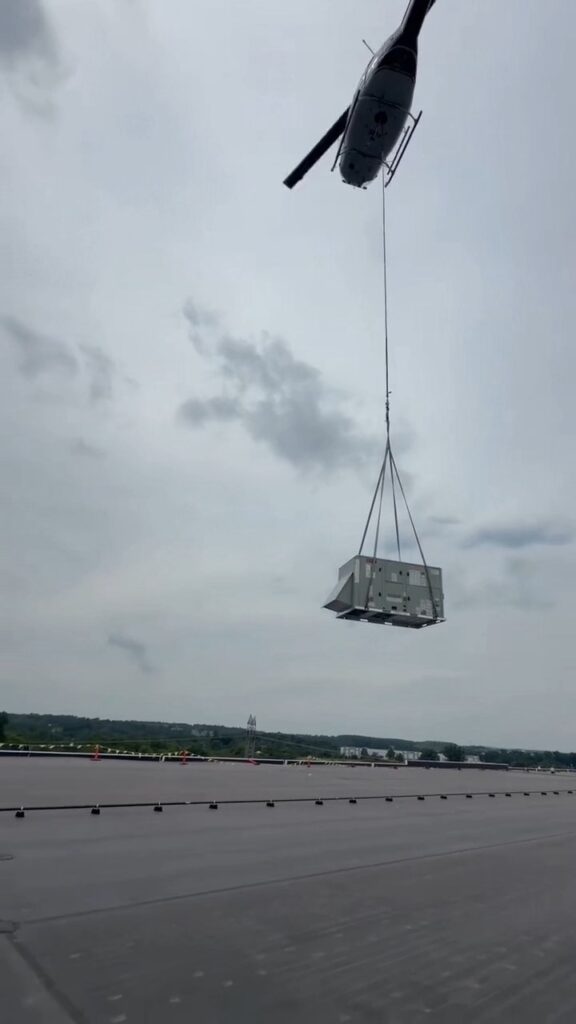 Helicopter lowering a new packaged rooftop HVAC unit onto a warehouse roof, seen from the roof deck