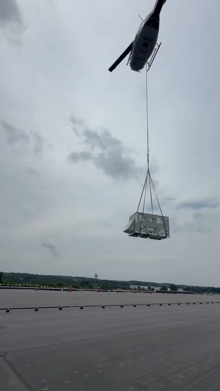 Helicopter lowering a new packaged rooftop HVAC unit onto a warehouse roof, seen from the roof deck