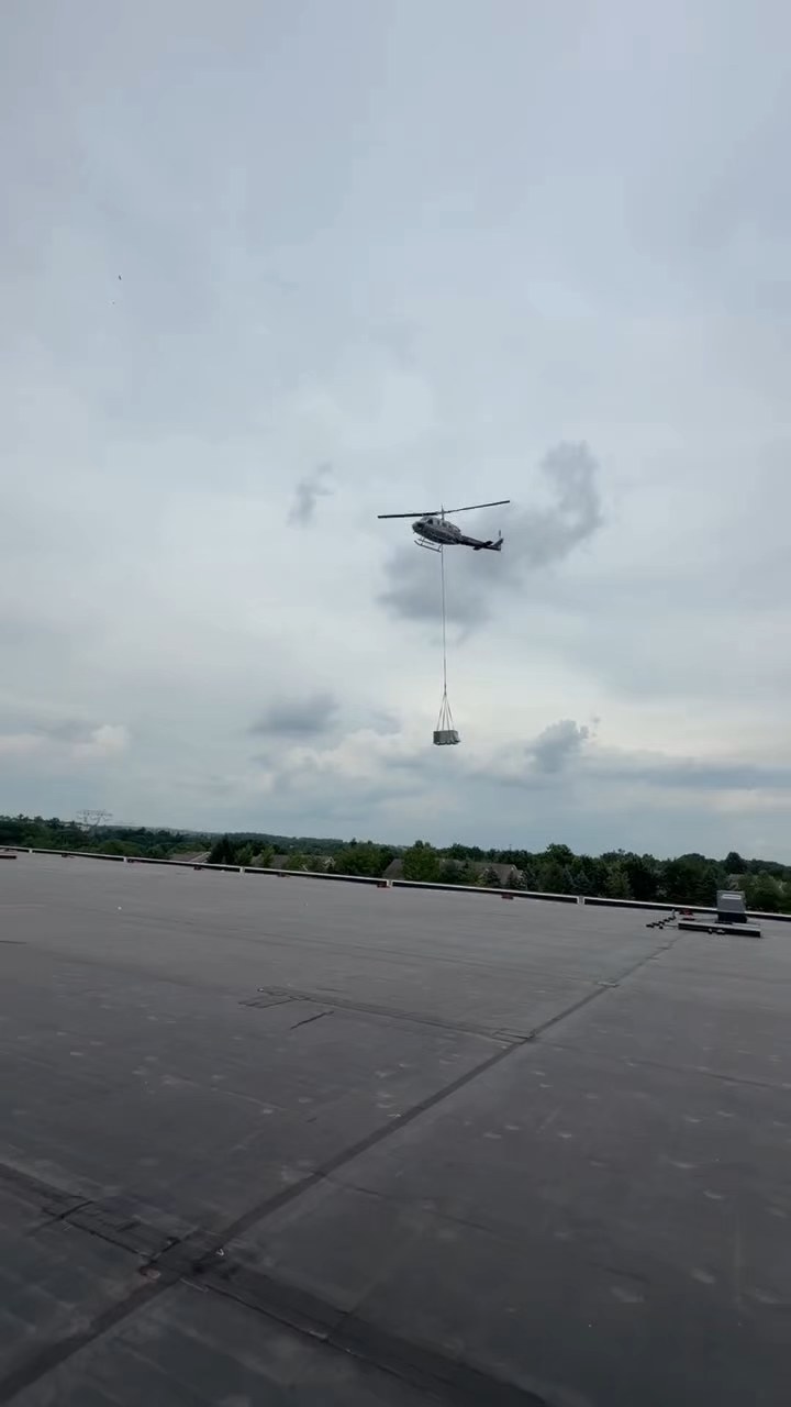 Wide view of a helicopter carrying a rooftop unit over a warehouse roof