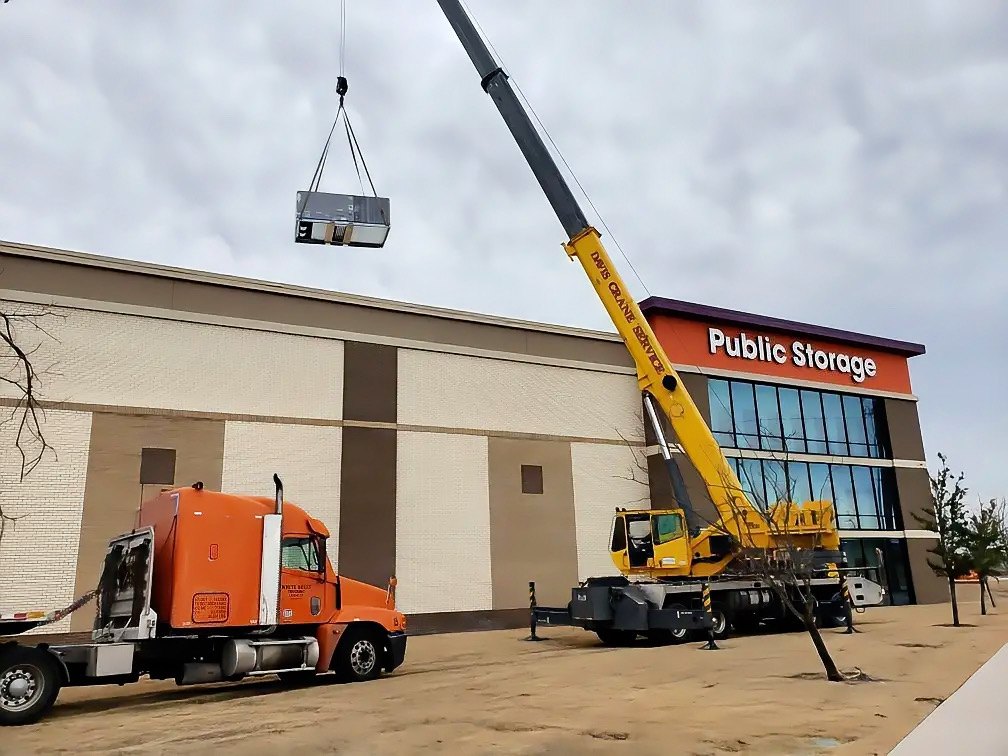 Crane lifting a packaged rooftop HVAC unit onto a new self-storage building, with the crane truck and tractor staged in front